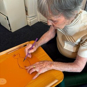 Elderly woman drawing at a table with colored pencils.
