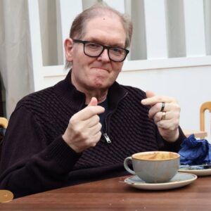 An older man smiling while sitting at a table with a cup of coffee.