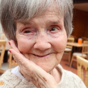 Close-up portrait of a smiling elderly woman with short grey hair, resting her face gently on her hand in a café setting.